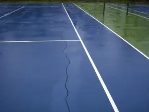 Cracked tennis court surface with visible line damage and water on outdoor court